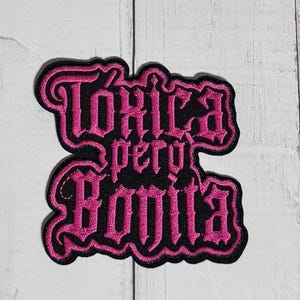Op de afbeelding: Een zwarte geborduurde patch met felroze letters die Tóxica pero Bonita spellen. De tekst is gerangschikt in drie regels in een gestileerd gotisch lettertype, met een dikke roze rand die de contouren van de letters volgt.