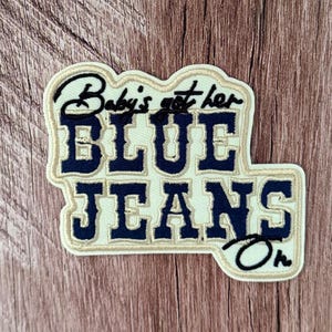 May include: An embroidered patch with a cream-colored background and dark navy blue lettering. The text reads Baby's got her BLUE JEANS On, featuring a mix of cursive and block fonts with a thick cream border outlining the entire shape of the patch.