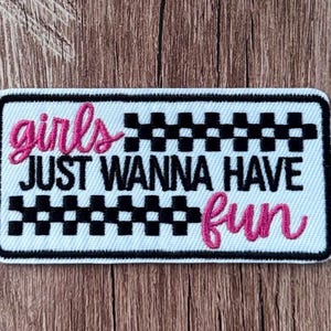 May include: A rectangular embroidered patch with a white background and a black border. It features the text "girls" in pink script, "JUST WANNA HAVE" in black block letters, and "fun" in pink script, accented by black and white checkered patterns.