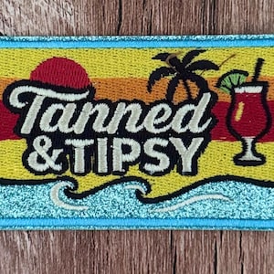 Puede incluir: Un parche bordado rectangular con un borde de purpurina azul claro. El diseño presenta un fondo de atardecer, la silueta de una palmera negra, una copa de cóctel roja con una rodaja de lima y el texto blanco Tanned & TIPSY sobre olas del mar.