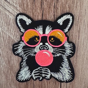 May include: An embroidered patch featuring a black and white raccoon wearing round pink-rimmed sunglasses with orange lenses. The raccoon is blowing a large, bright pink bubble with its mouth, and the design is rendered in detailed thread work.