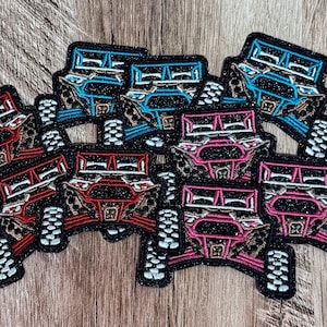 May include: A collection of embroidered patches shaped like off-road vehicles, featuring glittery black outlines and accents in bright blue, red, and pink. Each patch depicts the front view of a utility terrain vehicle with detailed tires.