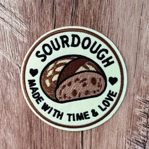 May include: A circular embroidered patch with a cream-colored background and a brown border. The center features a brown sourdough bread loaf illustration. The text reads SOURDOUGH MADE WITH TIME & LOVE, accented by two small black hearts.