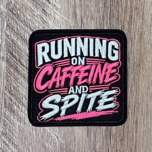 May include: A square embroidered patch with a black background featuring the white text RUNNING ON and SPITE, and the pink text CAFFEINE AND. The lettering has a distressed, brush-stroke style with a thick black border around the entire patch.