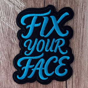 Puede incluir: Un parche bordado negro con el texto FIX YOUR FACE en hilo azul cian brillante. Las letras están en un estilo de escritura cursiva negrita y el parche tiene un borde troquelado que sigue la forma del texto, creando un contorno definido.