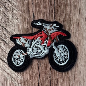 Pode incluir: Um emblema bordado em forma de motocicleta de motocross. O design apresenta um tanque de combustível e assento em vermelho brilhante, com detalhes do motor em cinza claro e branco, pneus pretos com raios brancos e uma borda preta espessa.