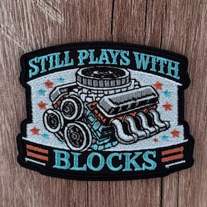 Puede incluir: Un parche bordado negro con una ilustración de motor en azul claro en el centro. El texto STILL PLAYS WITH está bordado en azul claro sobre el motor, y la palabra BLOCKS está bordada en azul claro debajo del motor.