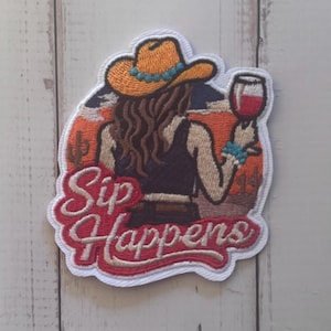 May include: An embroidered patch featuring a person wearing a yellow cowboy hat and a black top, holding a glass of red wine. The background shows a desert landscape with a cactus and the text Sip Happens in red script lettering with a white outline.