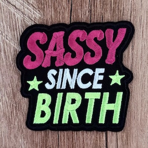 May include: A black embroidered patch featuring the text SASSY in pink, SINCE in white, and BIRTH in lime green. The design includes two lime green stars on either side of the word SINCE, all set against a black background with a black border.