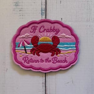 May include: An embroidered patch with a pink scalloped border featuring a red crab on a sandy beach with a yellow sun, blue ocean, and a pink umbrella. The text reads If Crabby Return to the Beach in dark pink thread against a light pink background.