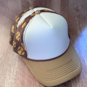 May include: A trucker hat featuring a solid white front panel, a tan brim with stitched detailing, and a brown mesh back panel patterned with yellow softball graphics. The cap includes a brown cord trim separating the front panel from the brim.