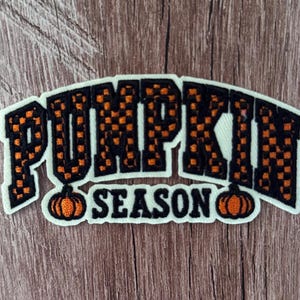Peut inclure: Un écusson brodé avec une bordure blanche présentant le texte PUMPKIN SEASON dans un motif à carreaux orange et noir. Le mot PUMPKIN est arqué au-dessus du mot SEASON, qui est flanqué de deux petites citrouilles brodées orange et noir.