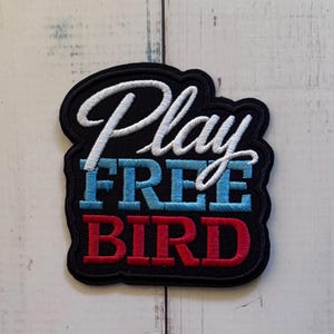 Könnte beinhalten: Ein schwarzer bestickter Aufnäher mit dem weißen kursiven Schriftzug Play, dem hellblauen Blocktext FREE und dem roten Blocktext BIRD. Der Aufnäher hat einen dicken schwarzen Rand und eine dem Textverlauf folgende Konturform.