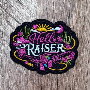 May include: An embroidered patch with a black background featuring the text Hell RAISER in pink and white. The design includes a pink cowboy hat, a revolver, cacti, roses, a yellow sun, and a crescent moon, all outlined in a scalloped black border.