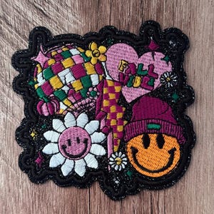 Pode incluir: Um emblema bordado com uma bola de discoteca, um coração rosa com o texto FALL VIBE, uma margarida com um rosto sorridente e um rosto sorridente usando um gorro roxo. O design inclui um raio xadrez amarelo e rosa e pequenas estrelas.