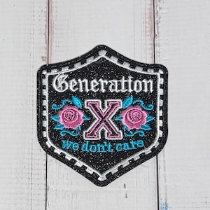 Puede incluir: Un parche bordado en forma de escudo con fondo negro brillante. Presenta el texto blanco Generation, una letra X grande de color rosa, dos rosas rosas con hojas verde azulado y el texto verde azulado We don't care, bordeado por un ribete a cuadros blanco y negro.
