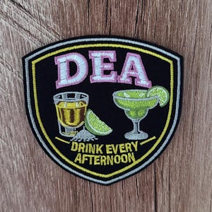 May include: A shield-shaped embroidered patch with a black background and a yellow and white border. It features the pink text DEA above a shot glass of yellow liquid, a lime wedge, salt, and a green margarita glass with the text DRINK EVERY AFTERNOON.