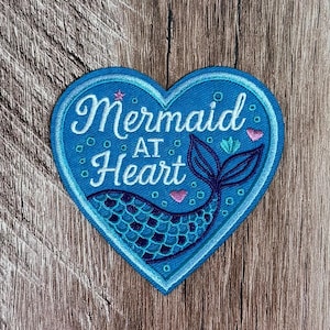Puede incluir: Un parche bordado en forma de corazón en tonos azules con un gráfico de cola de sirena y el texto blanco Mermaid AT Heart. El diseño incluye pequeños corazones rosas, una estrella rosa diminuta y burbujas azul claro bordadas en la tela.