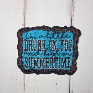 May include: A rectangular embroidered patch with a dark brown border and a bright blue background. The patch features black embroidered text that reads: I'm a little DRUNK ON YOU and high on SUMMERTIME in a mix of cursive and block lettering.