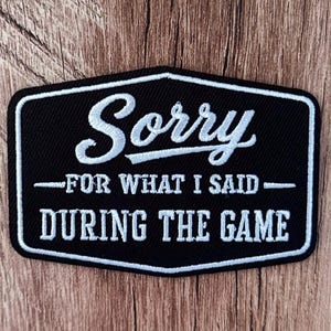 Può includere: Una toppa ricamata nera con bordo bianco e testo bianco che recita Sorry FOR WHAT I SAID DURING THE GAME. La toppa ha una forma a scudo con angoli arrotondati e una superficie in tessuto testurizzato, con un mix di caratteri corsivi e stampatello.