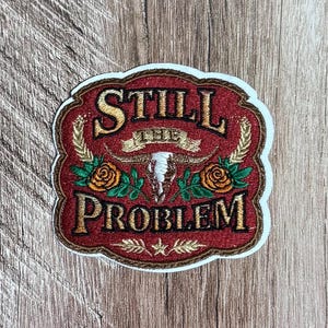 May include: An embroidered patch with a red background and a white border. The text reads STILL THE PROBLEM in gold lettering. The design features a central white steer skull, two orange roses with green leaves, and gold wheat stalks at the sides.