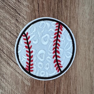 May include: A circular embroidered patch featuring a white baseball design with a subtle white leopard print pattern across the surface. The patch includes two curved red stitched seams and is bordered by a thick black embroidered edge.