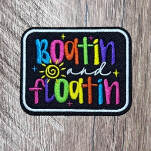 May include: A rectangular black embroidered patch with a white border. The text reads "Boatin and floatin" in multi-coloured, vibrant letters including pink, orange, green, blue, and yellow. A small yellow sun icon sits between the two lines.