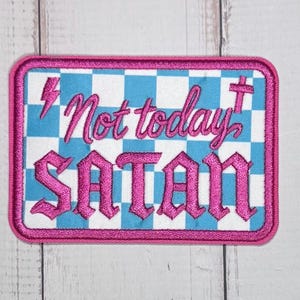 Pode incluir: Um emblema bordado retangular com um fundo xadrez azul claro e branco. O texto Not today está bordado em cursivo rosa acima da palavra SATAN em letras góticas rosa grandes. Um raio e uma cruz cor-de-rosa também estão incluídos.