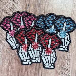 May include: A collection of embroidered patches featuring a white skeleton hand holding a finger to lips in a shushing gesture. The lips are patterned with a checkered design in red, pink, or blue, accented with a small flower at the wrist.