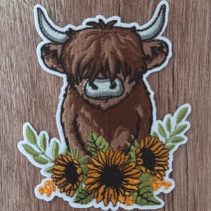 May include: An embroidered patch featuring a brown Highland cow with long curved horns, shaggy fur, and a light gray muzzle. The cow is surrounded by three yellow sunflowers with dark brown centers, green leaves, and small orange floral accents.
