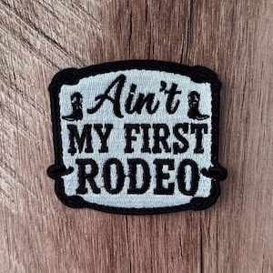 May include: A rectangular embroidered patch with a light gray background and a thick black border. The patch features the black text "Ain't MY FIRST RODEO" with a small black cowboy boot icon on each side of the word "Ain't" in a script font.