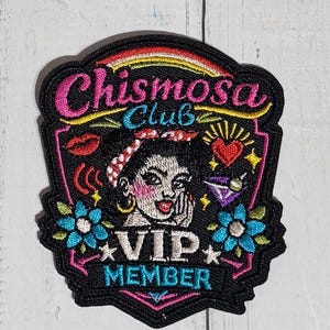 May include: A black embroidered patch featuring the text Chismosa Club and VIP MEMBER in pink and blue. The design includes a central illustration of a person with a red patterned headband, a red heart, a red lip icon, and blue flowers.