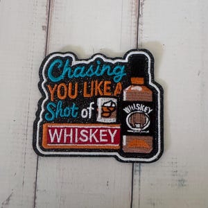 May include: An embroidered patch with a black glitter background featuring the text Chasing YOU LIKE A Shot of WHISKEY in teal, orange, and white thread. The design includes a small glass and a bottle of whiskey with a white label and barrel icon.