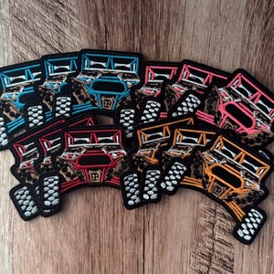 May include: A collection of embroidered patches shaped like off-road vehicles, featuring leopard print patterns on the body and black tires with white treads. The patches come in various frame colors including blue, pink, red, and orange.