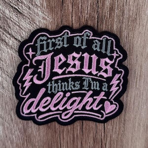 Op de afbeelding: Een zwarte geborduurde patch met roze en grijze letters die luidt, first of all Jesus thinks I'm a delight. Het ontwerp bevat twee roze bliksemschichten, twee kleine vierpuntige sterren en een klein roze hartje in de rechterbenedenhoek.