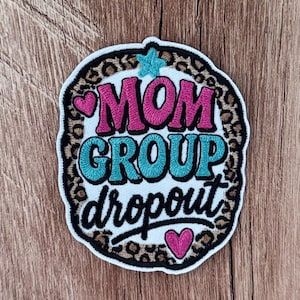 May include: An embroidered patch with a leopard print border and white center. The text reads MOM GROUP dropout in pink, teal, and black thread. A teal star sits at the top, with two pink hearts positioned on the left and bottom right sides.