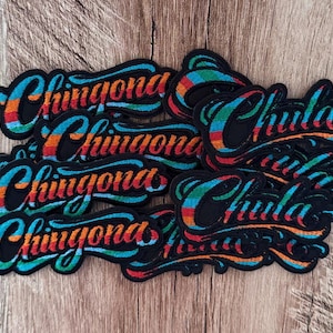 May include: A collection of embroidered patches featuring the words Chingona and Chula in a black script font. Each patch has a colorful border with horizontal stripes of red, orange, yellow, green, and blue thread, creating a vibrant effect.
