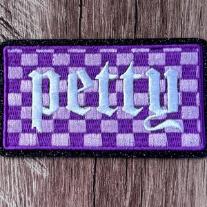 Puede incluir: Un parche rectangular bordado con un patrón de cuadros en violeta y violeta claro. La palabra petty está cosida en el centro con una fuente de estilo gótico en azul claro. El parche tiene un borde negro grueso con un acabado brillante.