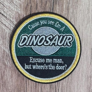 May include: A circular embroidered patch with a gold border. The center features a silver circle on a dark green background with the text 'Cause you see I'm A DINOSAUR' and a black lower section with the text 'Excuse me man, but where's the door?'