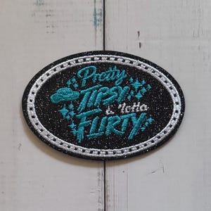 Puede incluir: Un parche bordado ovalado de color negro con purpurina que presenta el texto en color turquesa Pretty Tipsy & lotta FLIRTY. El diseño incluye pequeños detalles de estrellas bordadas y un borde blanco cosido alrededor del parche.