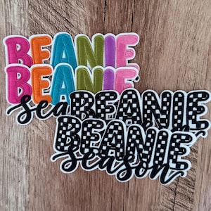 Pode incluir: Uma coleção de emblemas bordados com a palavra BEANIE em letras multicoloridas incluindo rosa, laranja, azul, verde e roxo, ao lado de emblemas xadrez preto e branco com o texto BEANIE SEASON em uma fonte de escrita preta.