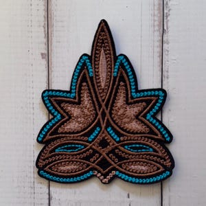 May include: An embroidered patch featuring a symmetrical, pointed design with brown and tan thread detailing. The pattern is outlined and accented with bright turquoise stitching against a black background, creating a sharp, decorative shape.