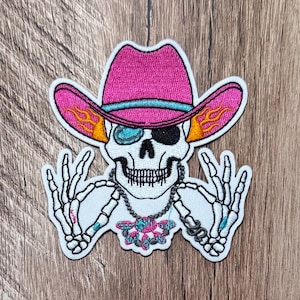 May include: An embroidered patch featuring a white skull wearing a bright pink cowboy hat with a turquoise band and orange flame accents. The skull has one turquoise eye and one black eye, wears a beaded necklace, and has two skeleton hands making signs.