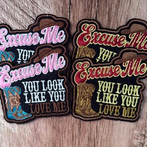 Peut inclure: Quatre écussons brodés noirs représentant un chapeau de cowboy marron, une botte de cowboy et le texte "Excuse Me YOU LOOK LIKE YOU LOVE ME". Deux écussons utilisent du fil rose et bleu, tandis que les deux autres utilisent du fil rouge et jaune.