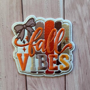 Pode incluir: Um emblema bordado com o texto fall VIBES em fio laranja e dourado. O design inclui um laço castanho no canto superior esquerdo, pequenos detalhes de estrelas e riscas verticais em tons de castanho, laranja e bronze atrás das letras.
