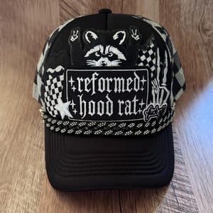 Op de afbeelding: Een zwarte truckerpet met een centrale patch met de tekst 'reformed hood rat' in witte gotische letters. Het ontwerp bevat een wasbeer-afbeelding, een witte ster, een skeletthand en een zwart-wit geblokt patroon op de zijpanelen.