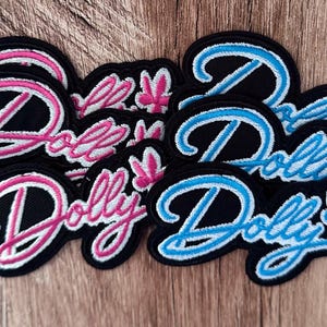 May include: A collection of embroidered patches featuring the word Dolly in cursive script. Some patches have bright pink lettering with a matching pink flower icon, while others have light blue lettering with a matching blue flower icon.