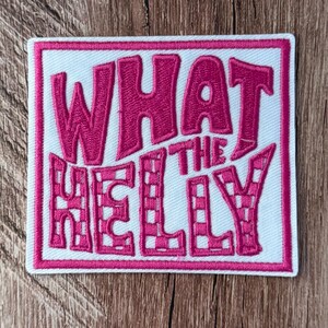 Puede incluir: Un parche bordado cuadrado con fondo blanco y borde rosa. El parche presenta el texto WHAT THE HELLY en letras rosas, negritas y estilizadas. Las letras H, E, L, L e Y tienen un patrón de cuadros dentro del bordado.