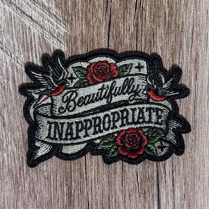 May include: An embroidered patch featuring a light gray banner with the black text "Beautifully INAPPROPRIATE". The design includes two black swallows with red accents, two red roses with green leaves, and a thick black border surrounding the shape.