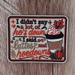 May include: A rectangular embroidered patch with a light background and dark border. It features the text 'I didn't say a lot of ho's down I said lattes and hoedowns' in brown and orange. An illustration of a coffee cup with whipped cream, a maple leaf, and a small pumpkin are included.